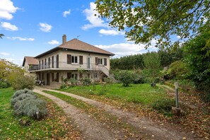 A two-story house with a balcony, surrounded by a garden and a dirt path.