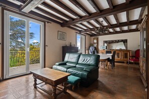 A spacious living area with a green leather sofa, wooden table, and a dining table with chairs. There is a balcony with a view of trees and a clear sky.