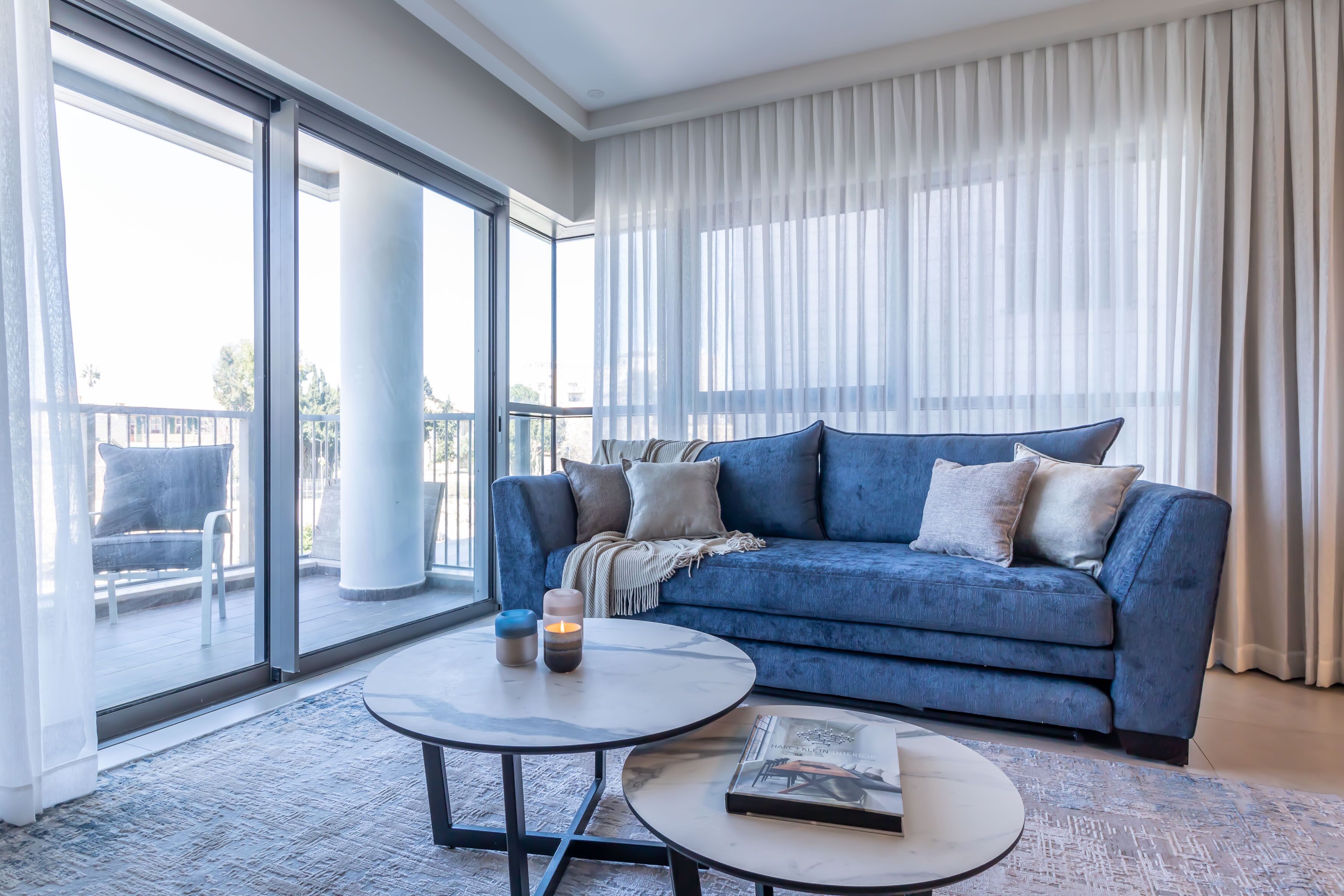 A modern living room with a blue sofa, two round coffee tables, and a large window with sheer curtains.