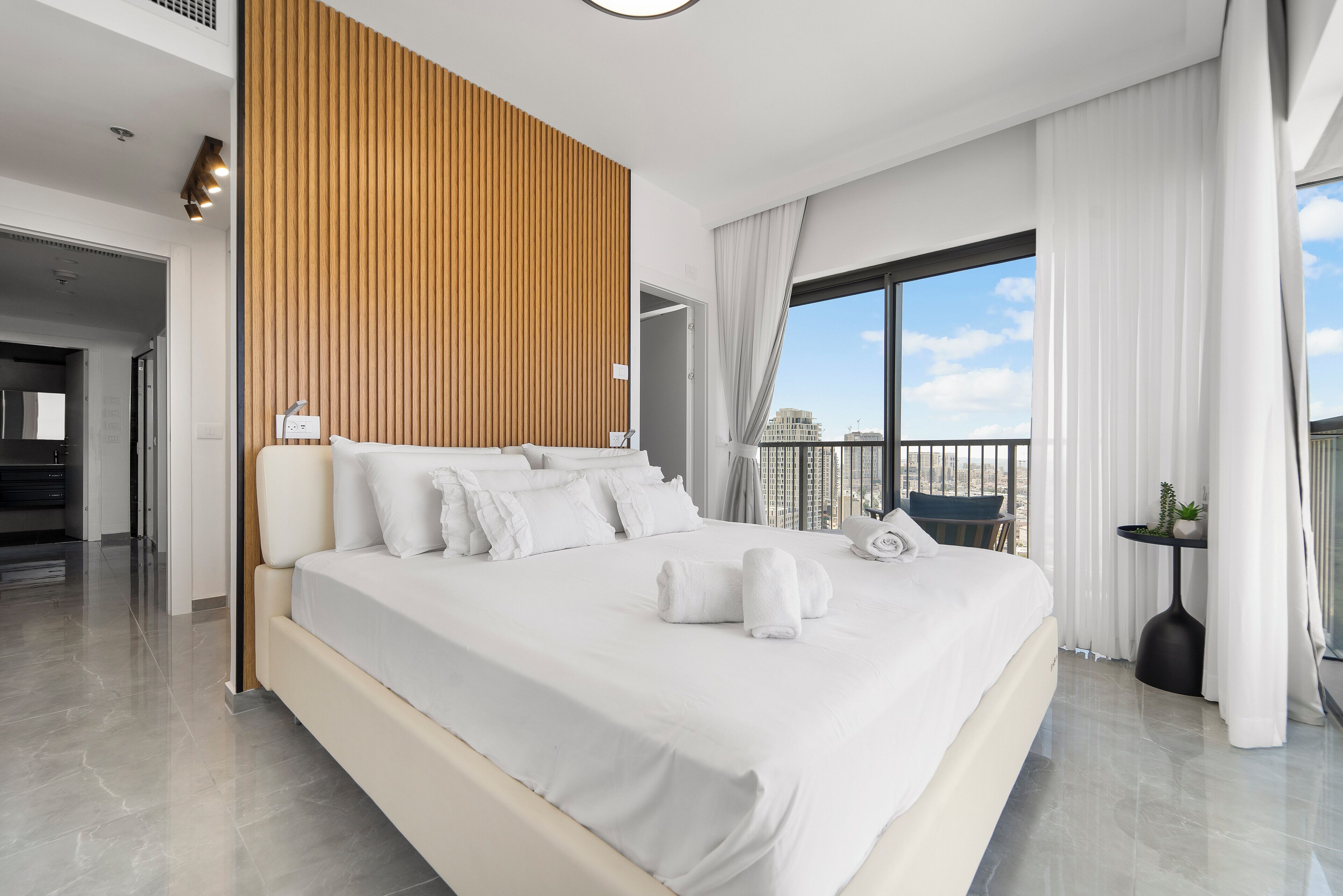 A modern hotel room with a large bed, a wooden accent wall, a balcony with a view, and a small table with a plant.