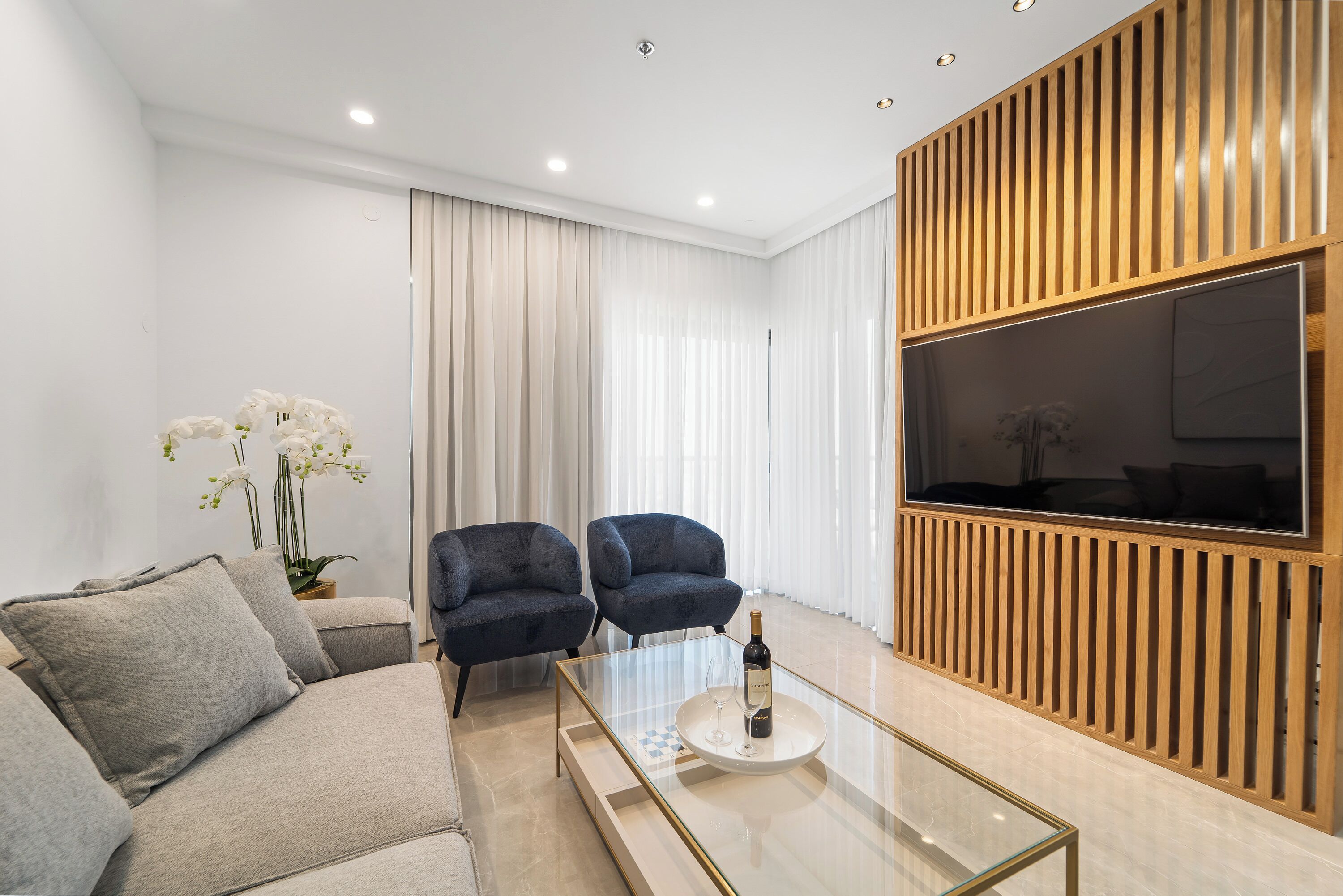 Un salon moderne avec un canapé, deux fauteuils, une table basse en verre sur laquelle se trouvent une bouteille et un verre, et un grand téléviseur fixé à un mur en bois.