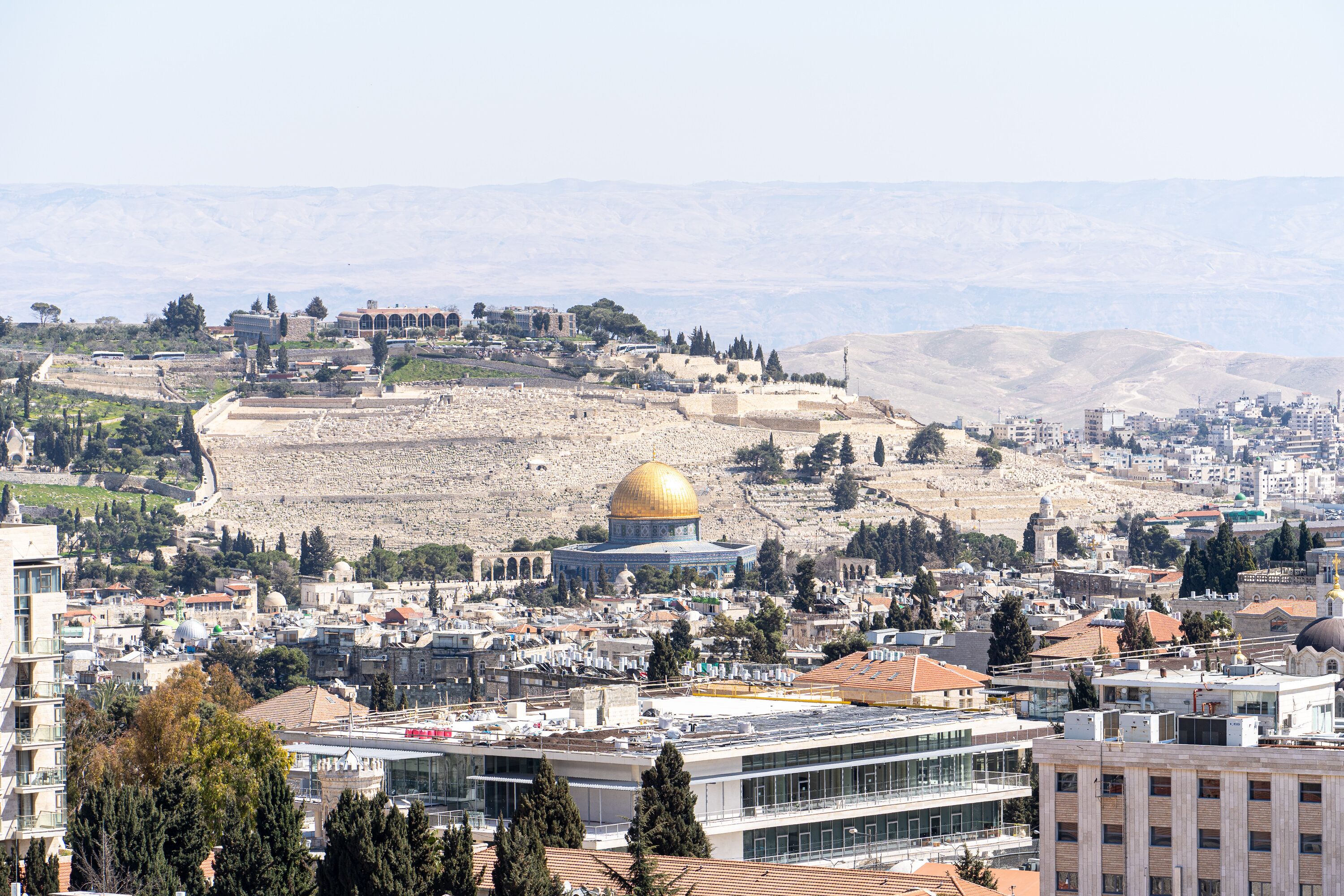 A cityscape with a prominent golden dome structure, modern buildings, and a hilly backdrop.