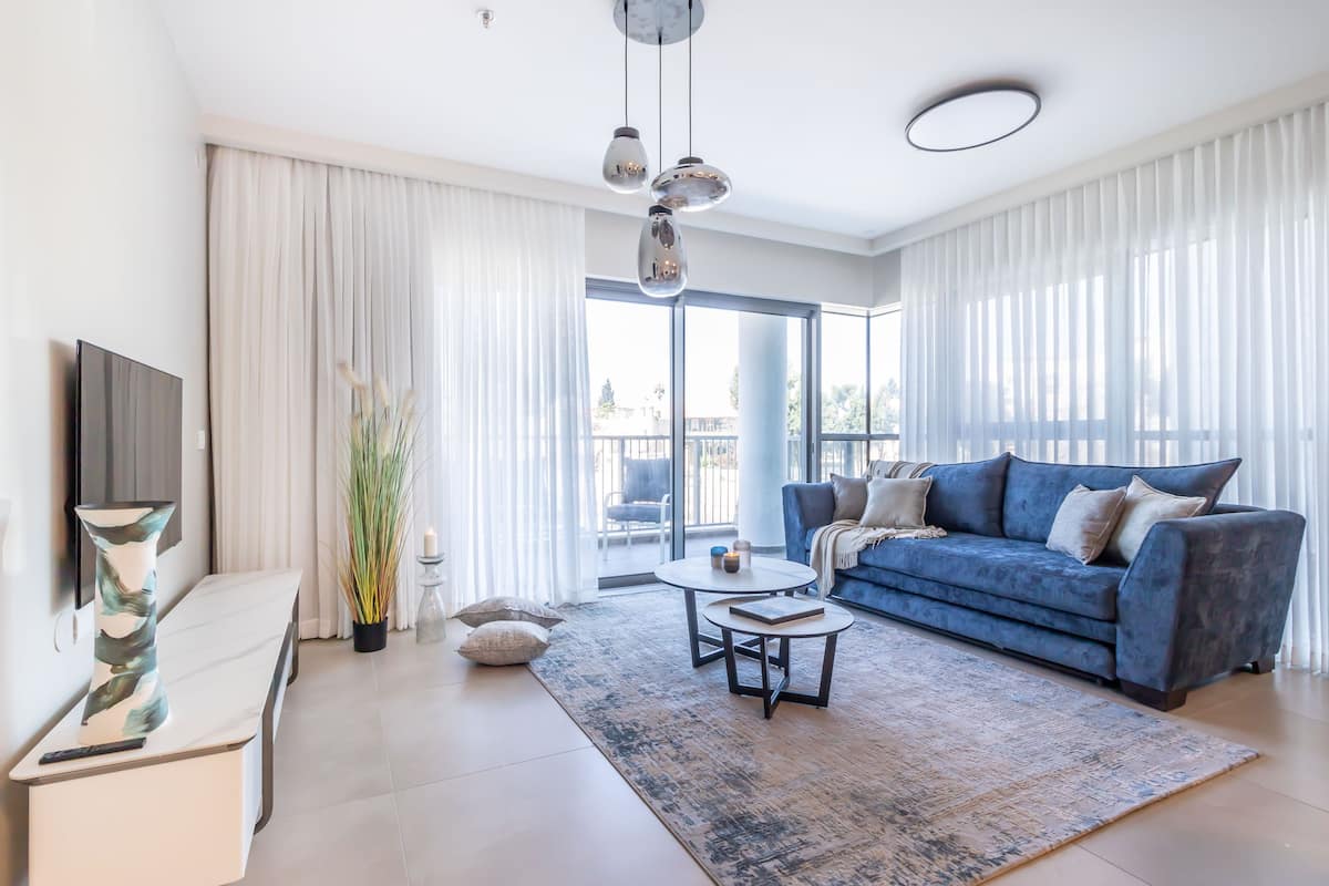 Un salon moderne avec un canapé bleu, une table basse blanche et une grande fenêtre ornée de rideaux légers.