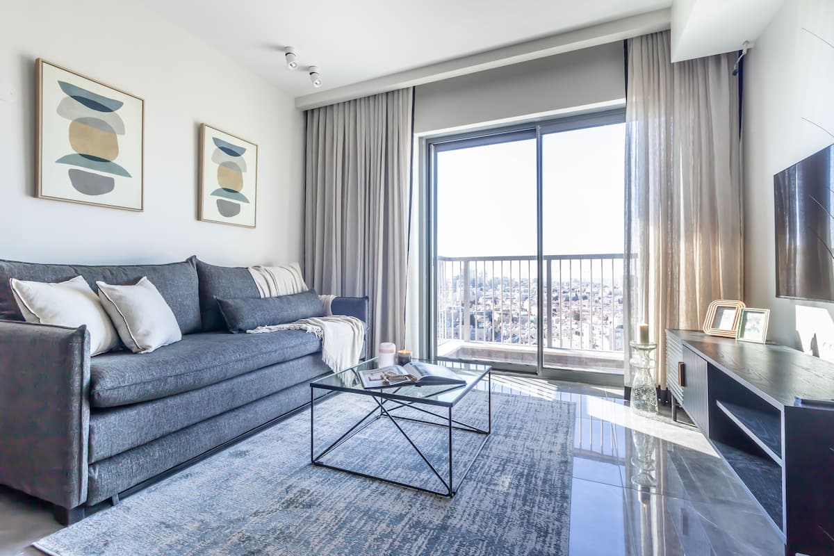 Un salon moderne avec un canapé gris, une table basse en verre et une grande fenêtre avec des rideaux.
