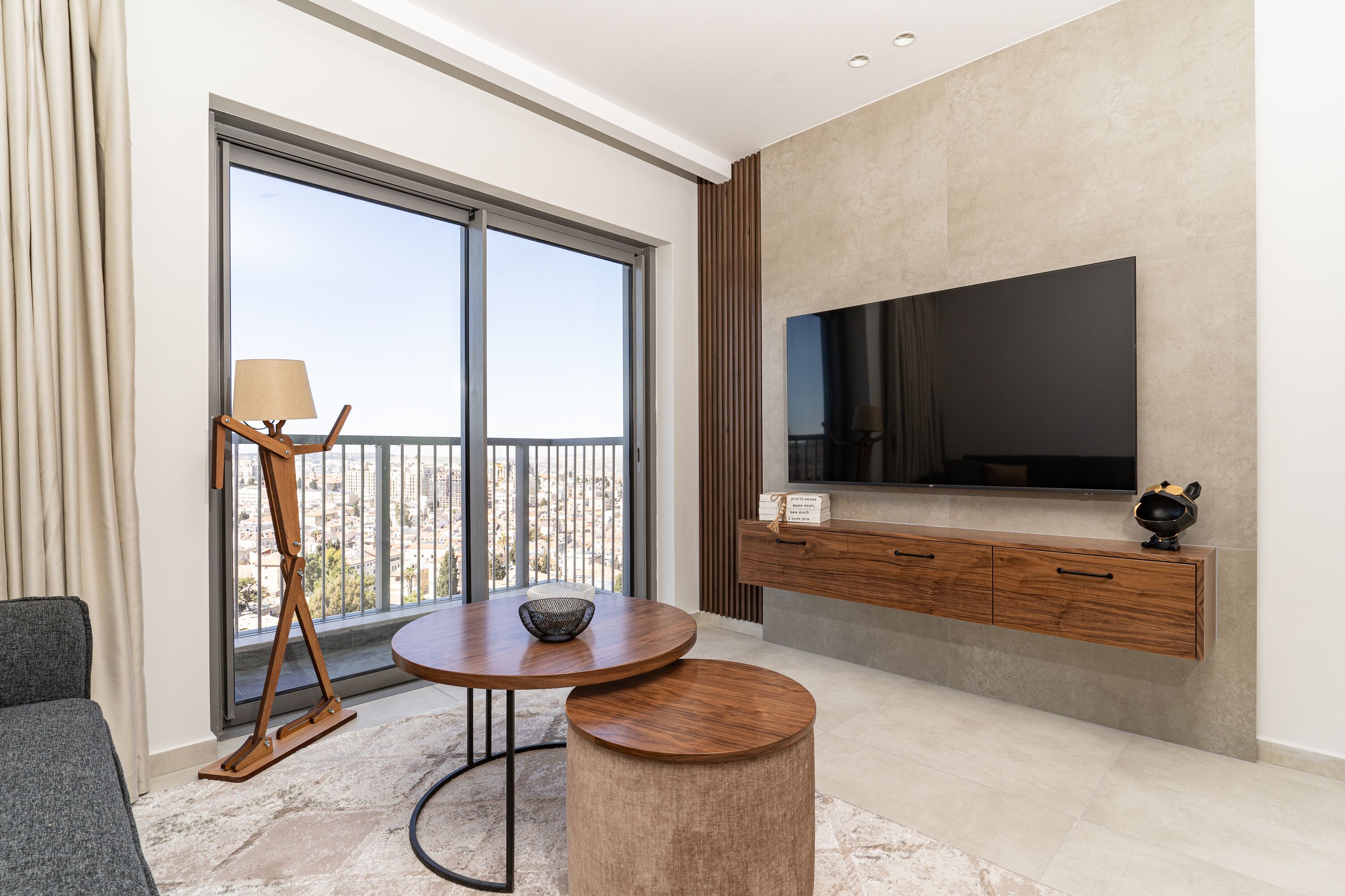 A modern living room with a large window, a flat-screen TV, a wooden cabinet, and a round table with a lamp.