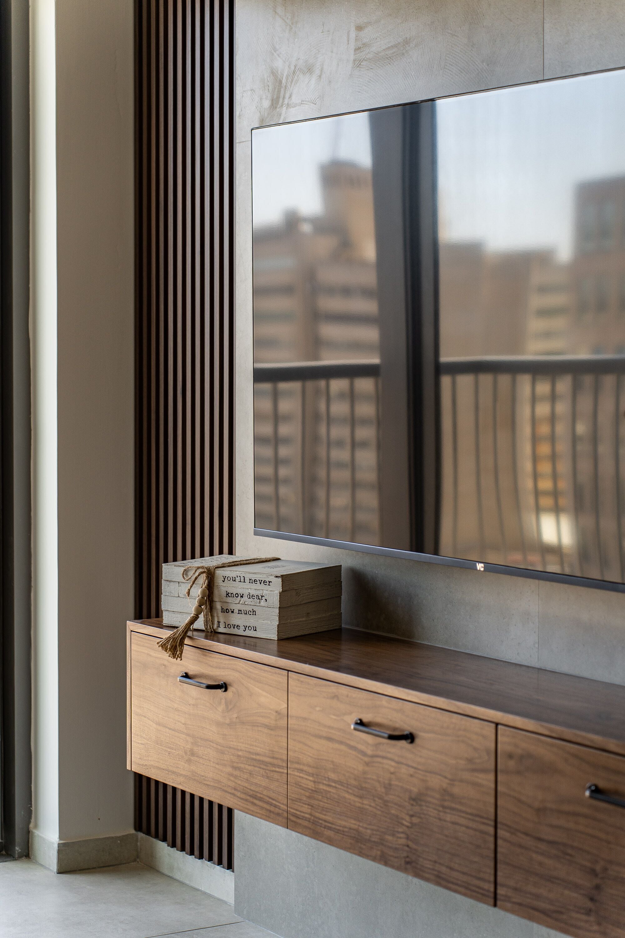 A modern living room with a large TV mounted on a wall, a wooden cabinet with drawers, and a window with a view of buildings.