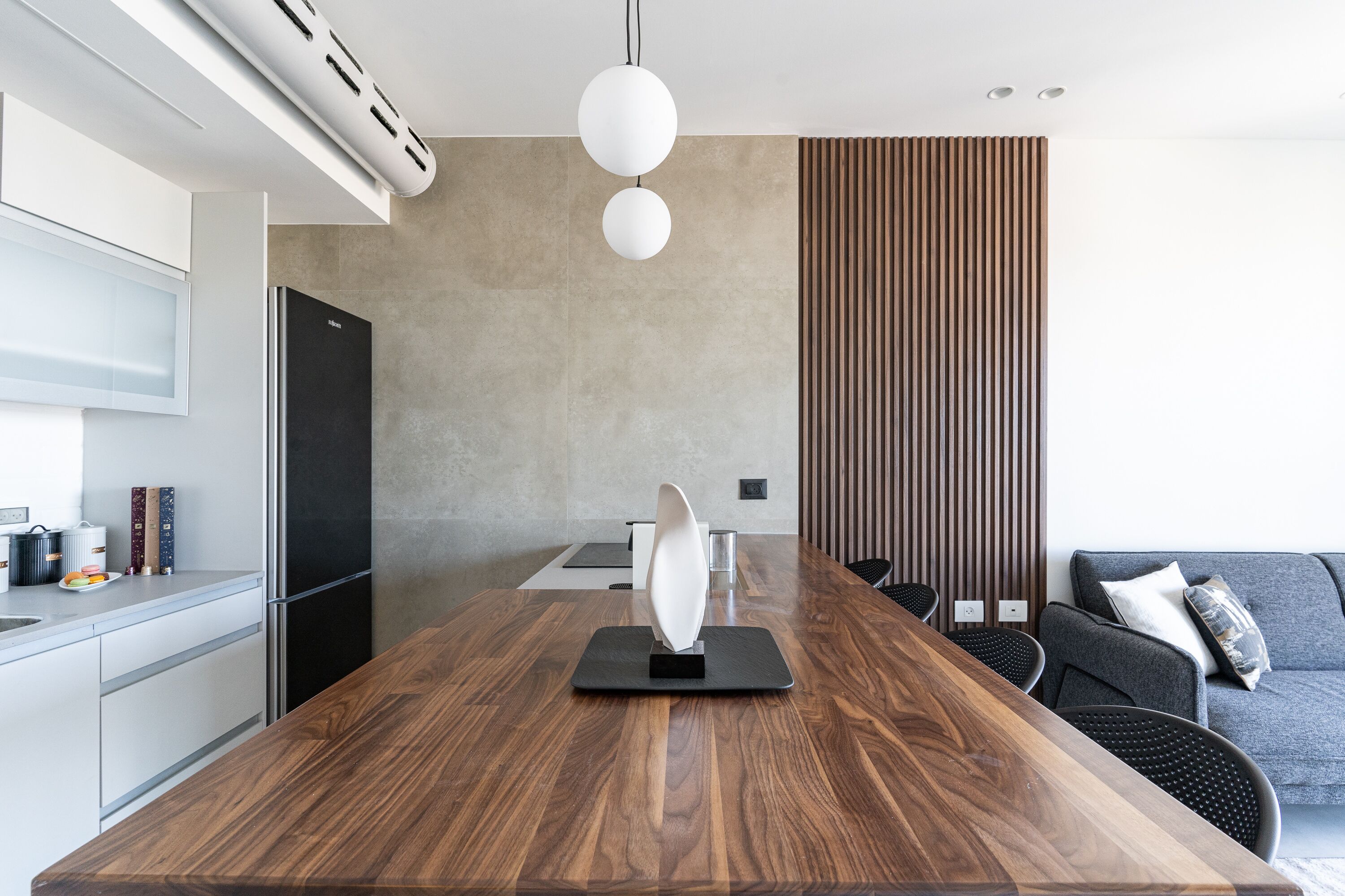 A modern kitchen with a wooden dining table, a black refrigerator, and a grey sofa in the background.