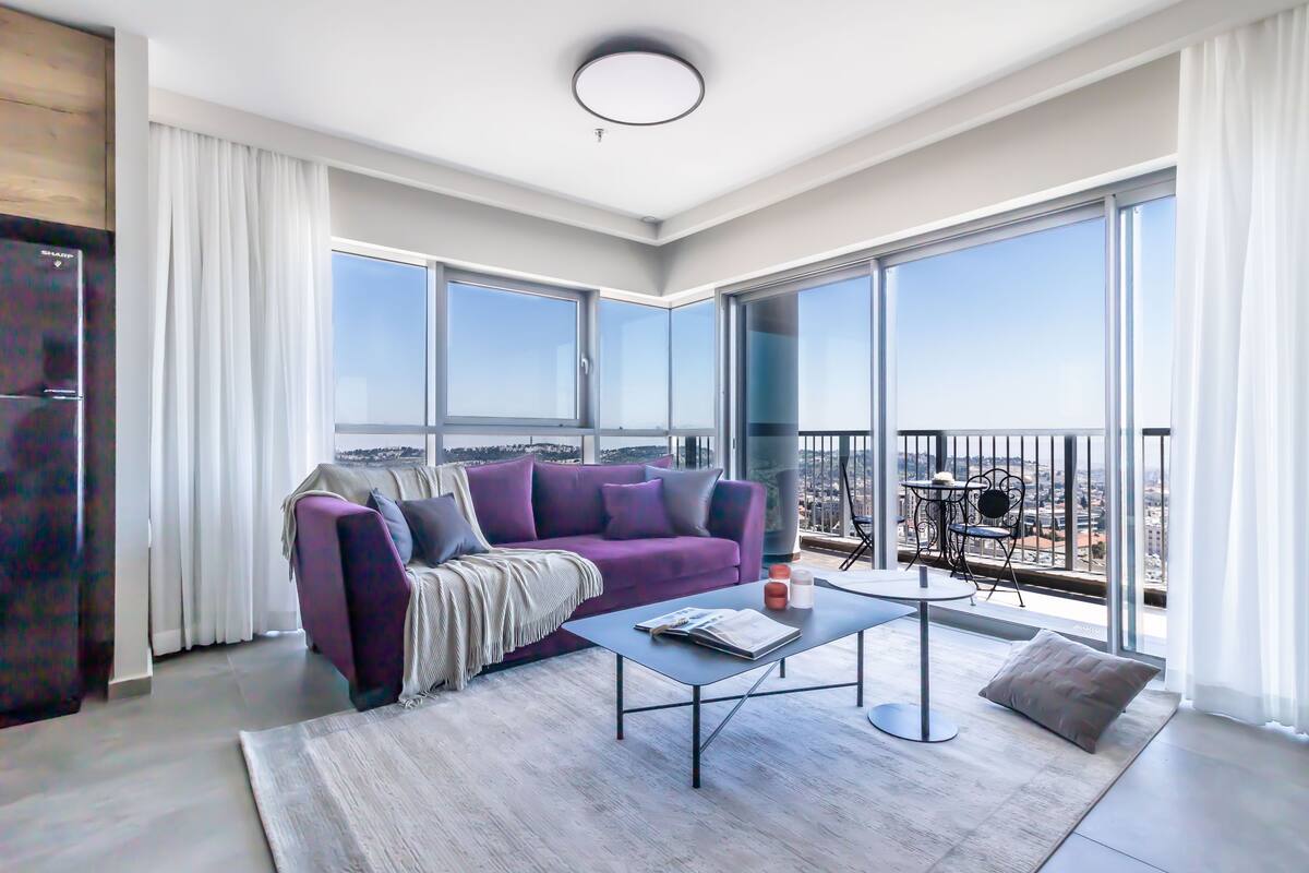 Un salon moderne avec un canapé violet, une table basse et un balcon offrant une vue sur la ville.