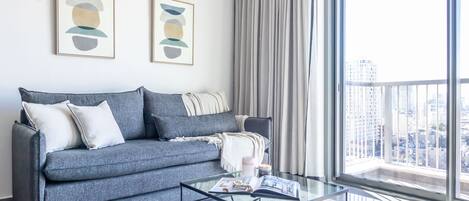 Un salon moderne avec un canapé gris, une table basse en verre et une grande fenêtre donnant sur la ville.