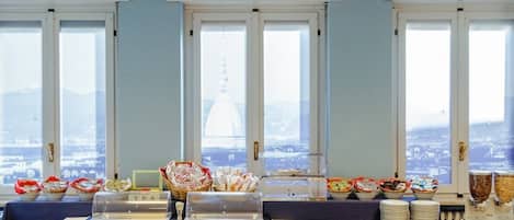 A buffet setup with various food items and a tea station, with a view of a cityscape through large windows.