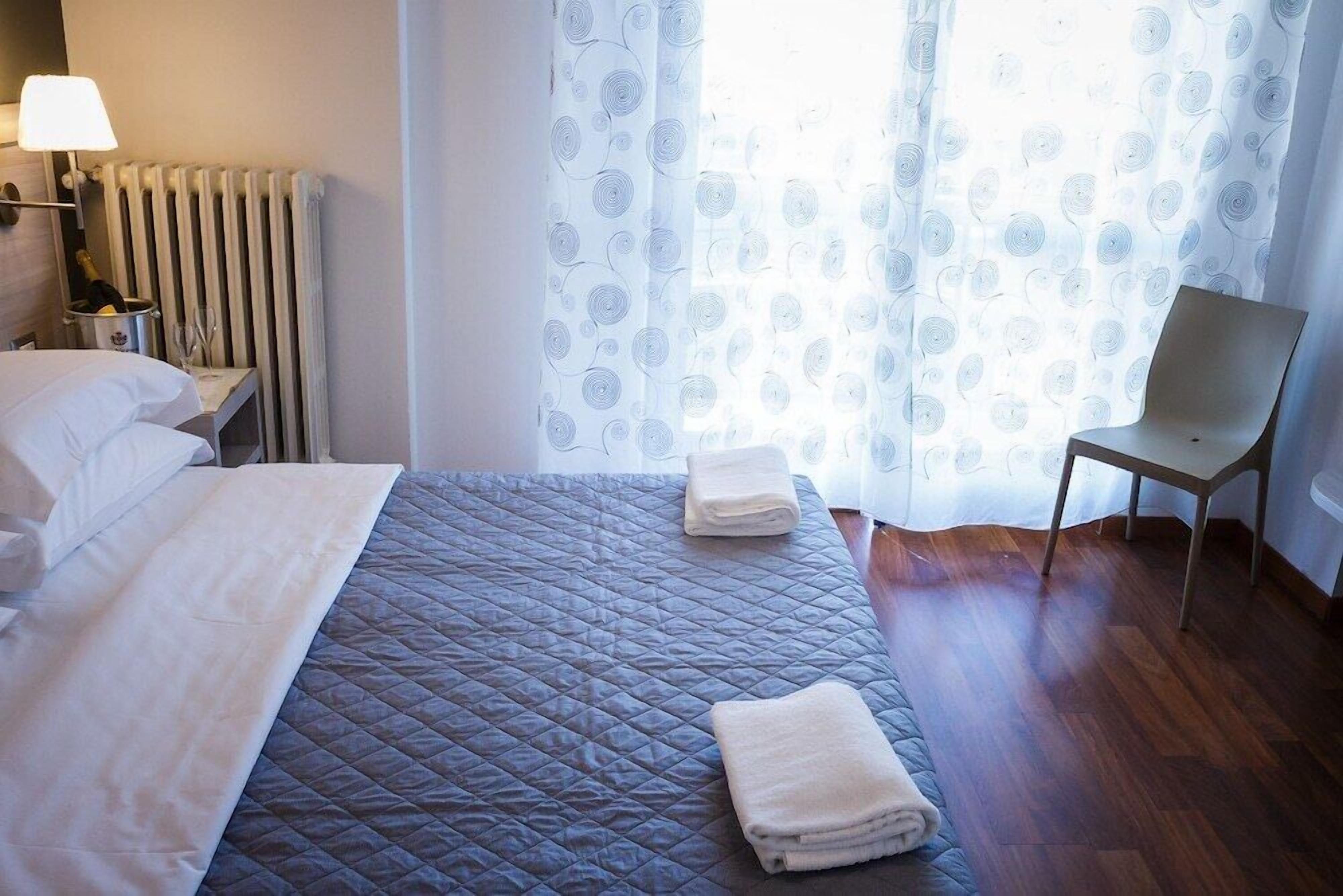 Una habitación de hotel con una cama, una silla, una lámpara, un radiador y una ventana con cortinas.