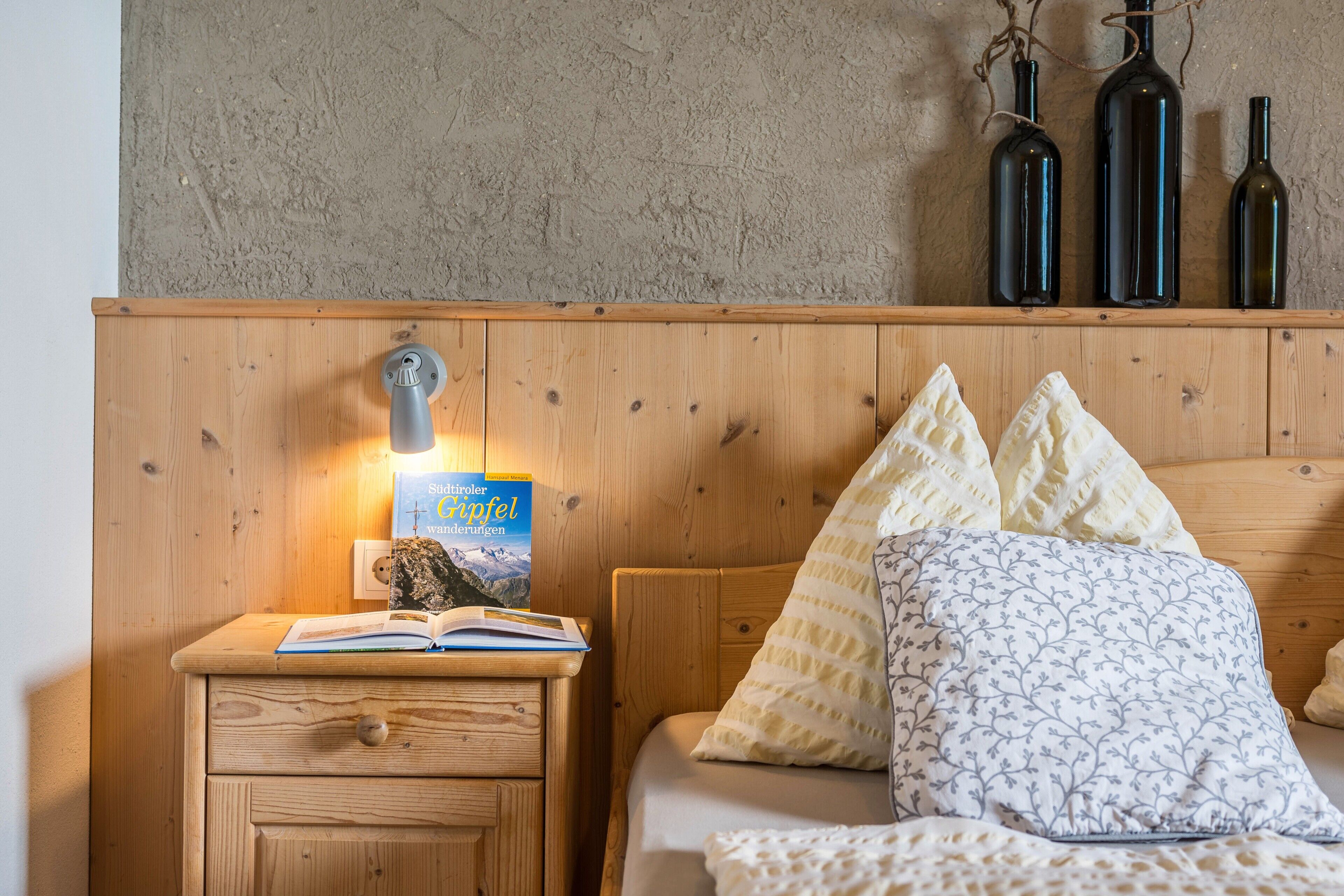 A wooden bedside table with a book and a lamp, a bed with patterned pillows, and three wine bottles on a textured wall.