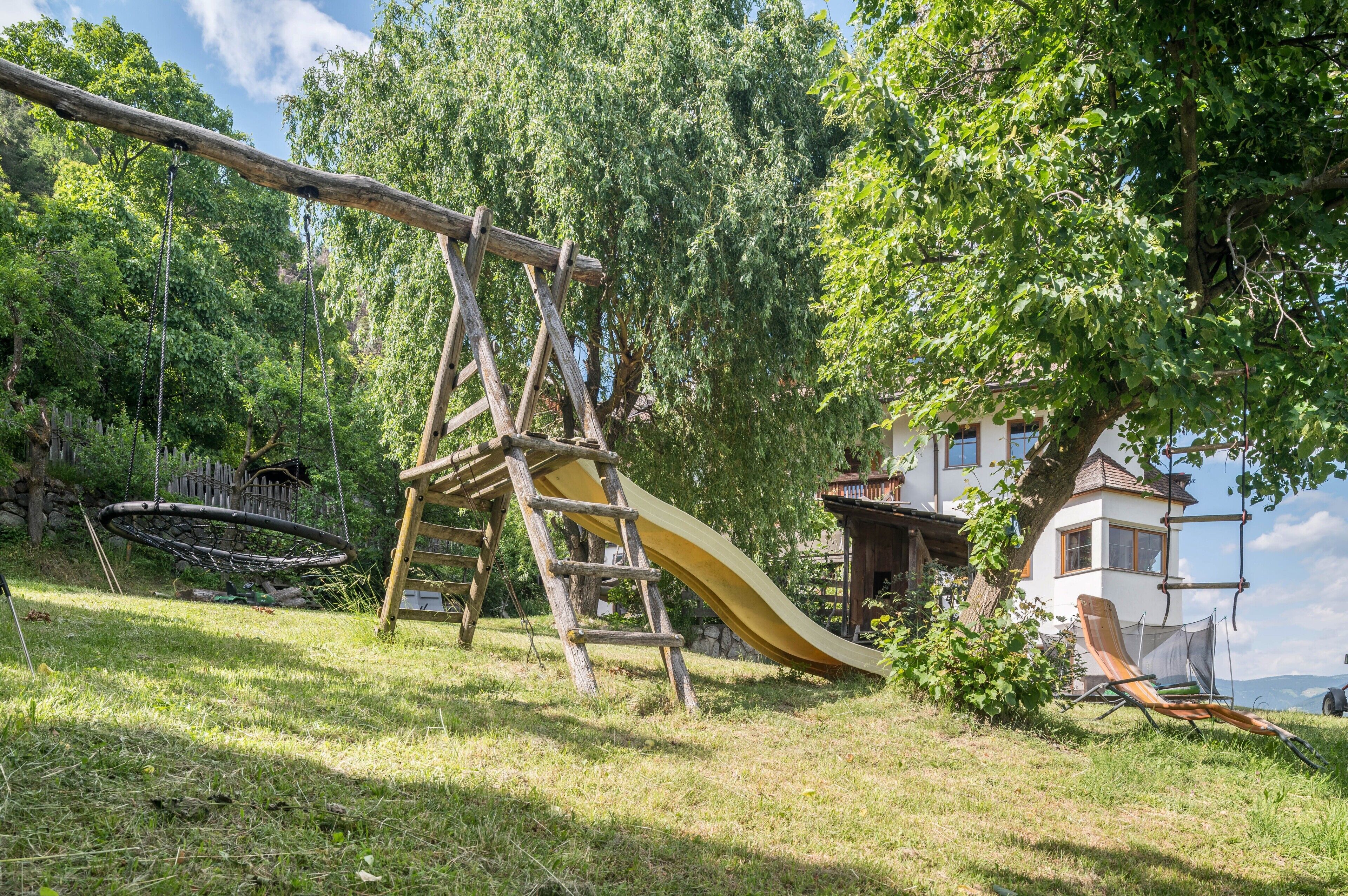 Una struttura di gioco in legno con uno scivolo, un'amaca e una casetta su un'area erbosa, con una casa sullo sfondo.