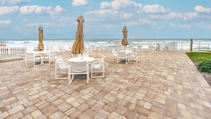 Un espace extérieur aménagé avec des tables et des chaises blanches, des parasols de couleur beige et une vue sur l’océan.