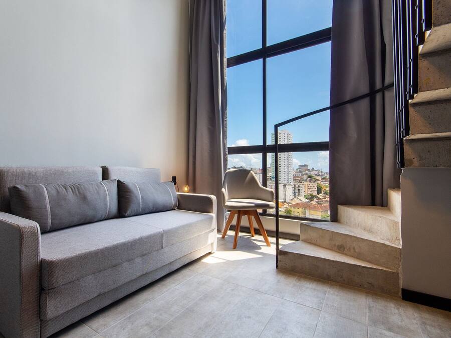 Una sala de estar moderna con un sofá gris, una silla de madera y una escalera que lleva a un balcón al aire libre.