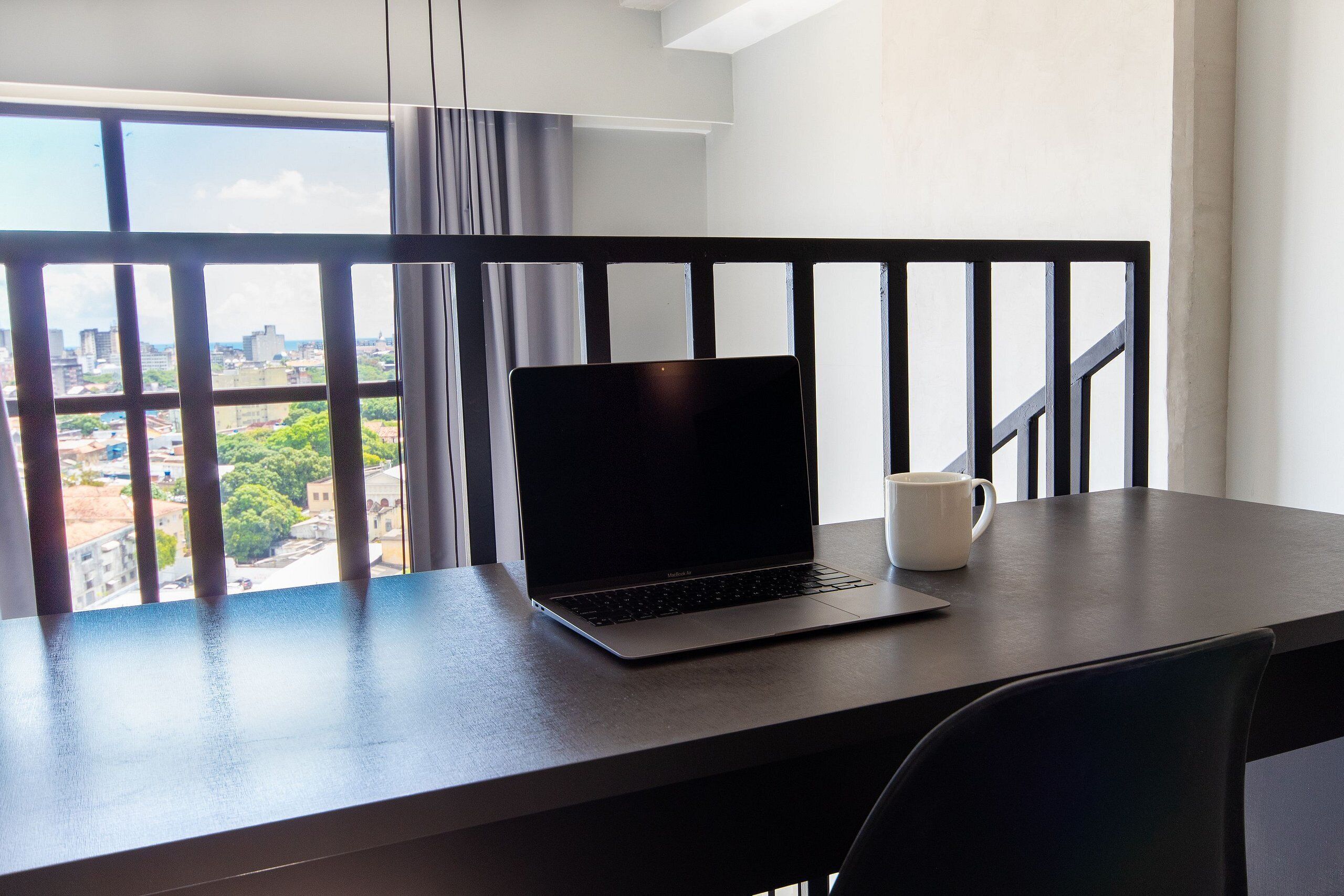 Un portátil sobre una mesa, con una taza blanca, y vistas de la ciudad a través de amplios ventanales y una barandilla negra.