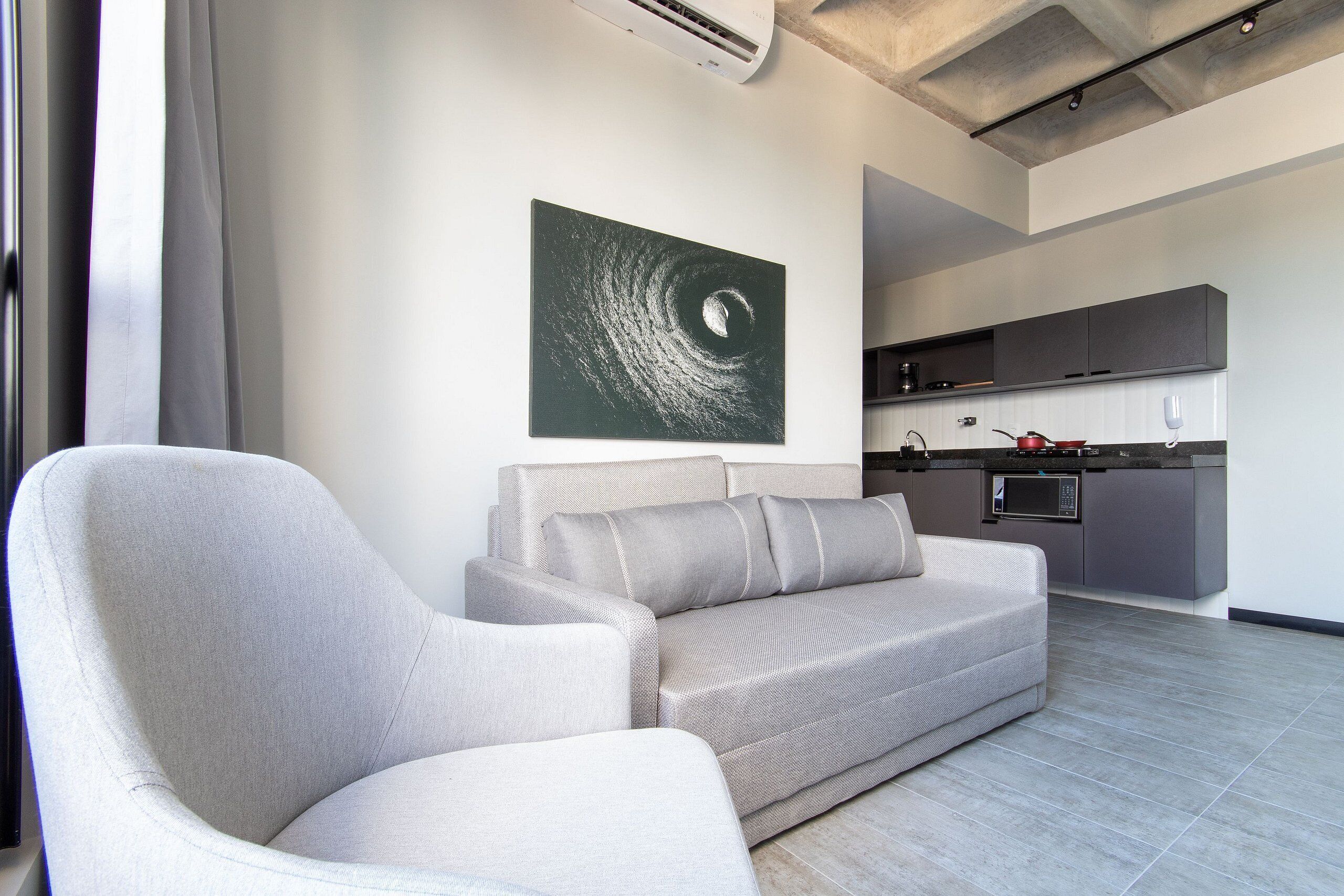 Una sala moderna con un sofá gris, un cuadro de un diseño ondulante y una cocina al fondo.