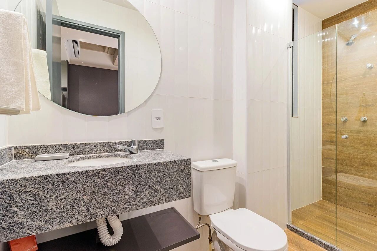 Uma casa de banho com bancada de granito, sanita, cabine de duche em vidro e espelho com porta-toalhas.