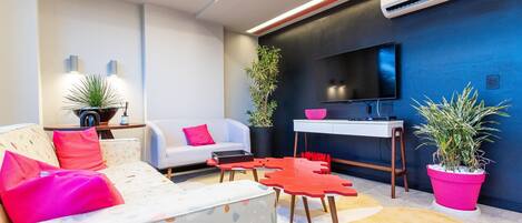 A modern living room with a sofa, coffee table, and TV. There are potted plants and a desk with a chair.