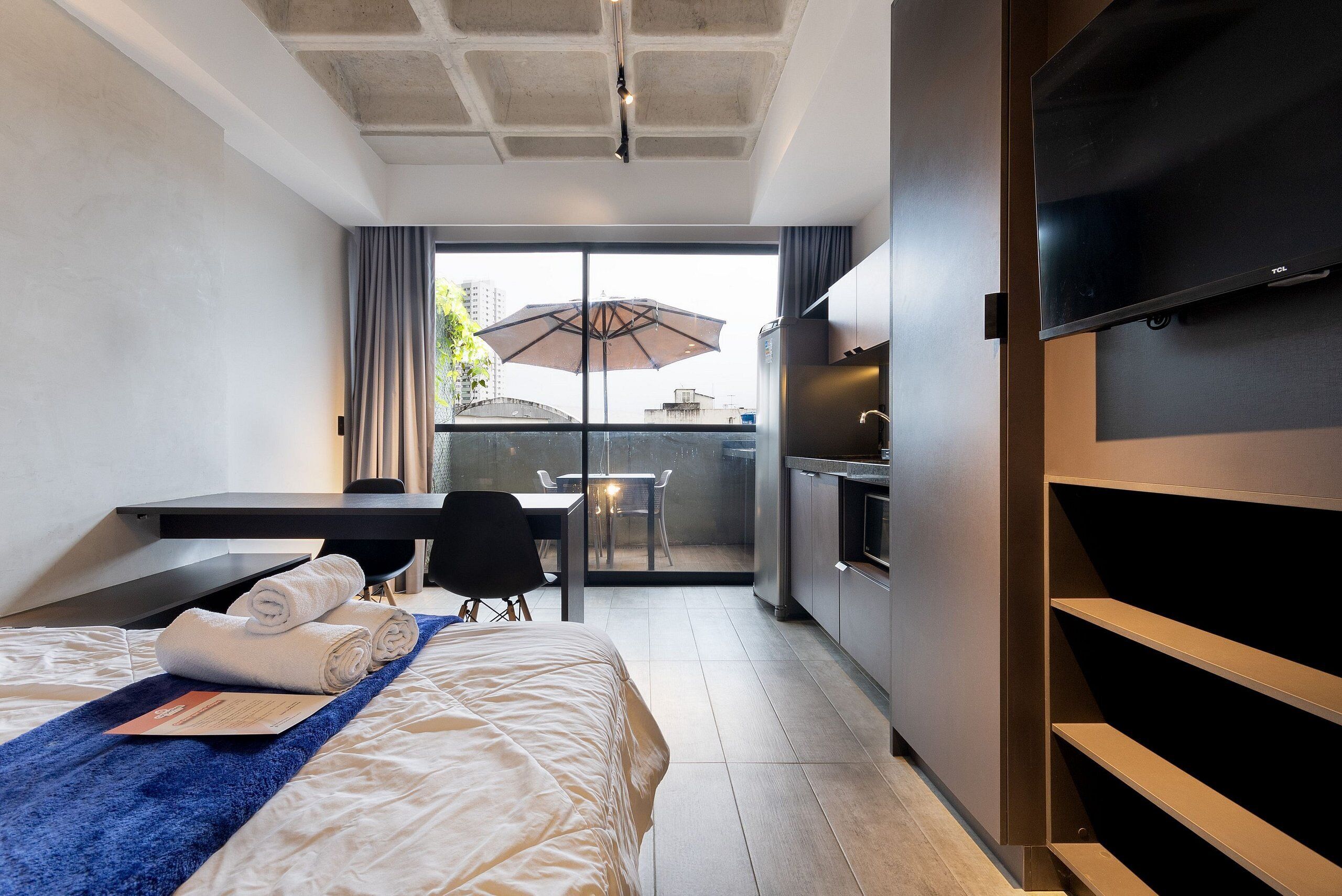 A hotel room with a bed, a desk, a chair, a TV, a balcony with an umbrella, and a kitchenette.