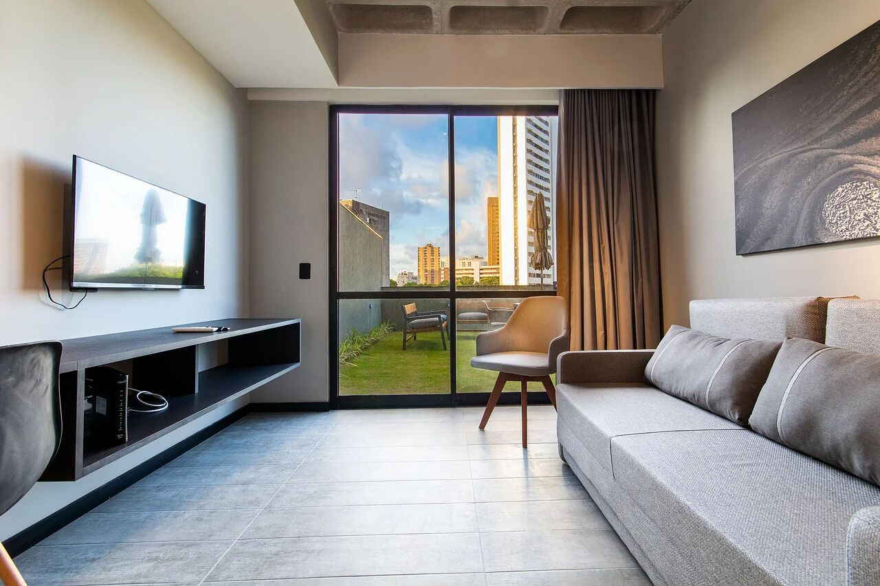 A modern living room with a flat-screen TV, a sofa, a chair, and a large sliding glass door leading to a balcony.