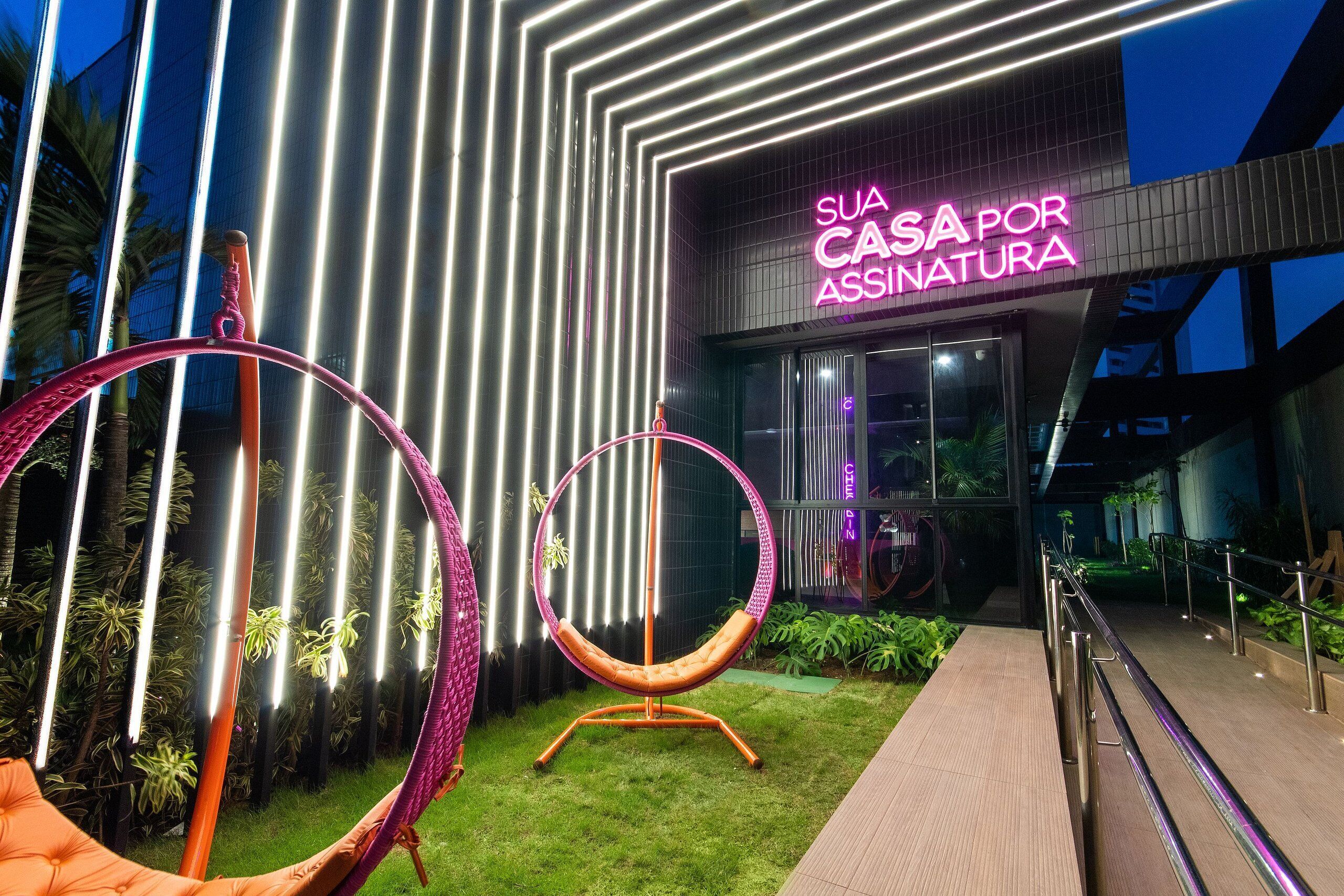 A modern building with a neon sign reading 'SUA CASA POR ASSINATURA' and two hanging chairs in the foreground.