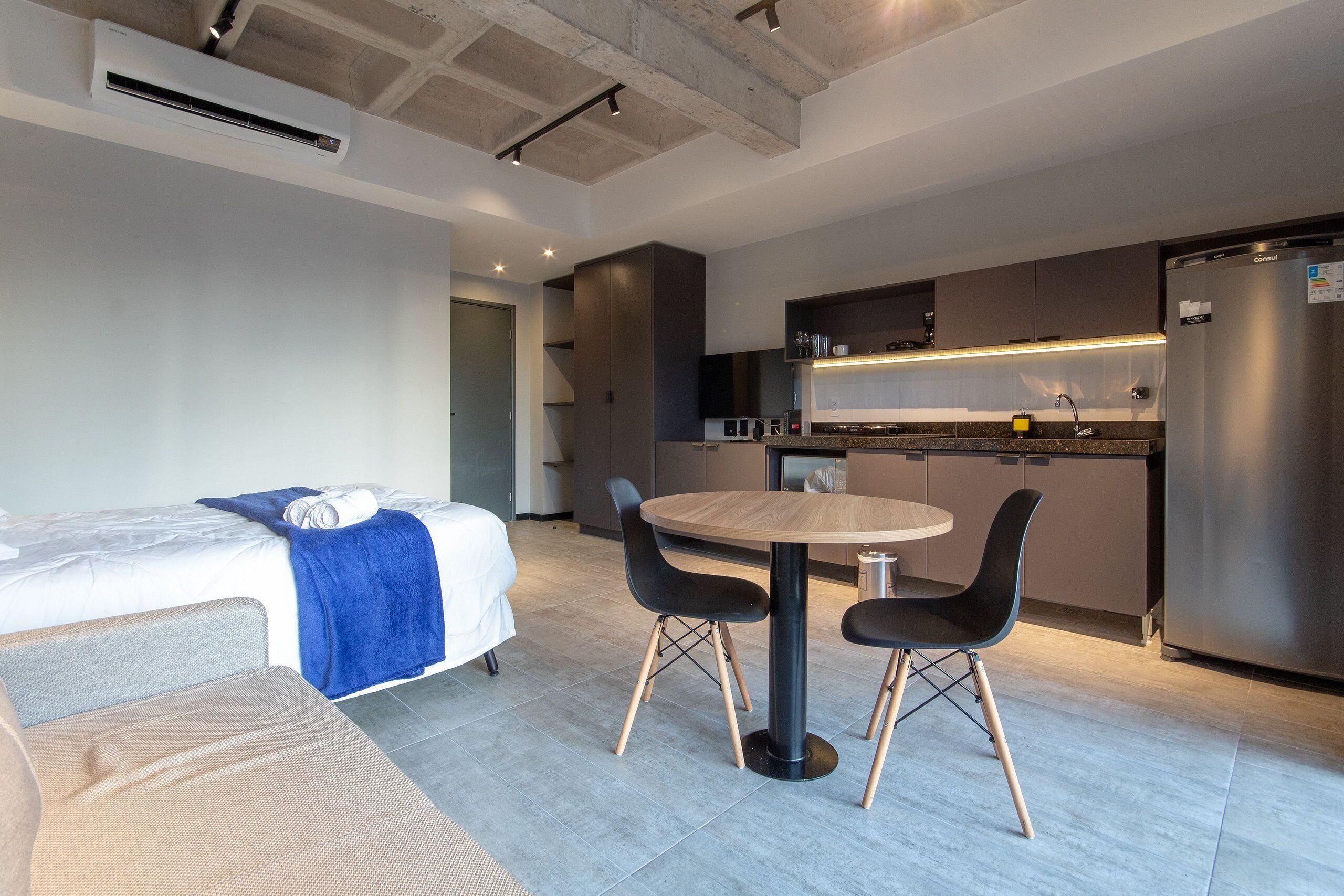 A modern hotel room with a bed, a sofa, a dining table with chairs, a kitchen area, and a built-in air conditioning unit.