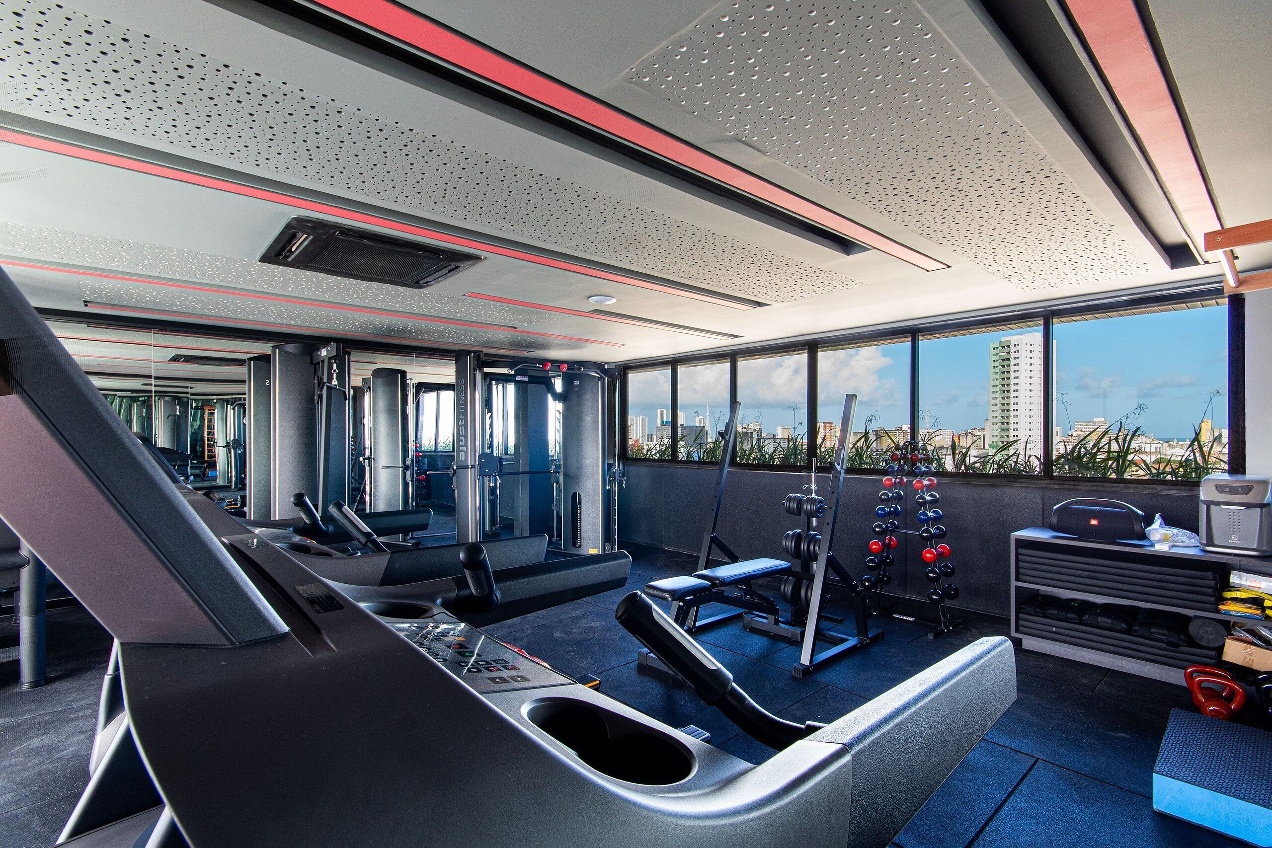 Un gimnasio con equipamiento de ejercicio, ventanas amplias y vista a la ciudad.