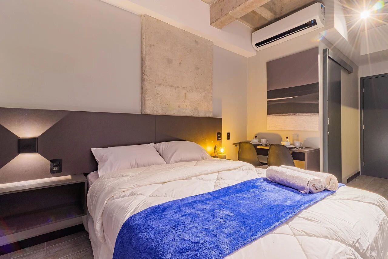 Moderni hotellihuone, jossa on sänky, yöpöytä, lamppu ja työpöytä tuolilla.