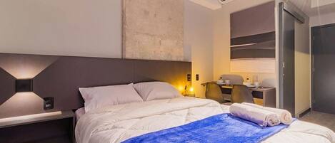 Moderni hotellihuone, jossa on sänky, yöpöytä, lamppu ja työpöytä tuolilla.