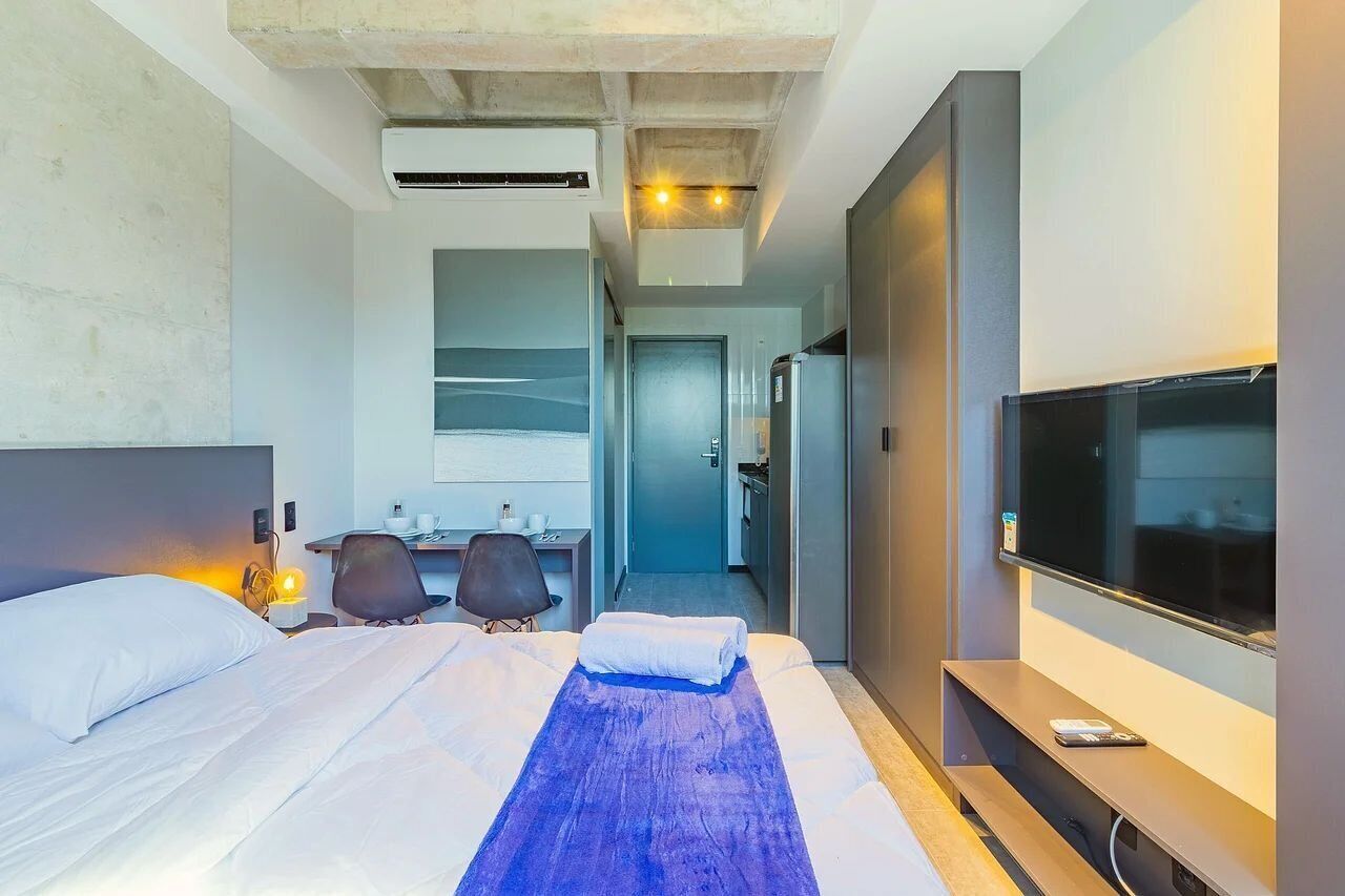 Une chambre d’hôtel avec un lit, un bureau, une chaise, une télévision et, au fond, une petite cuisine.