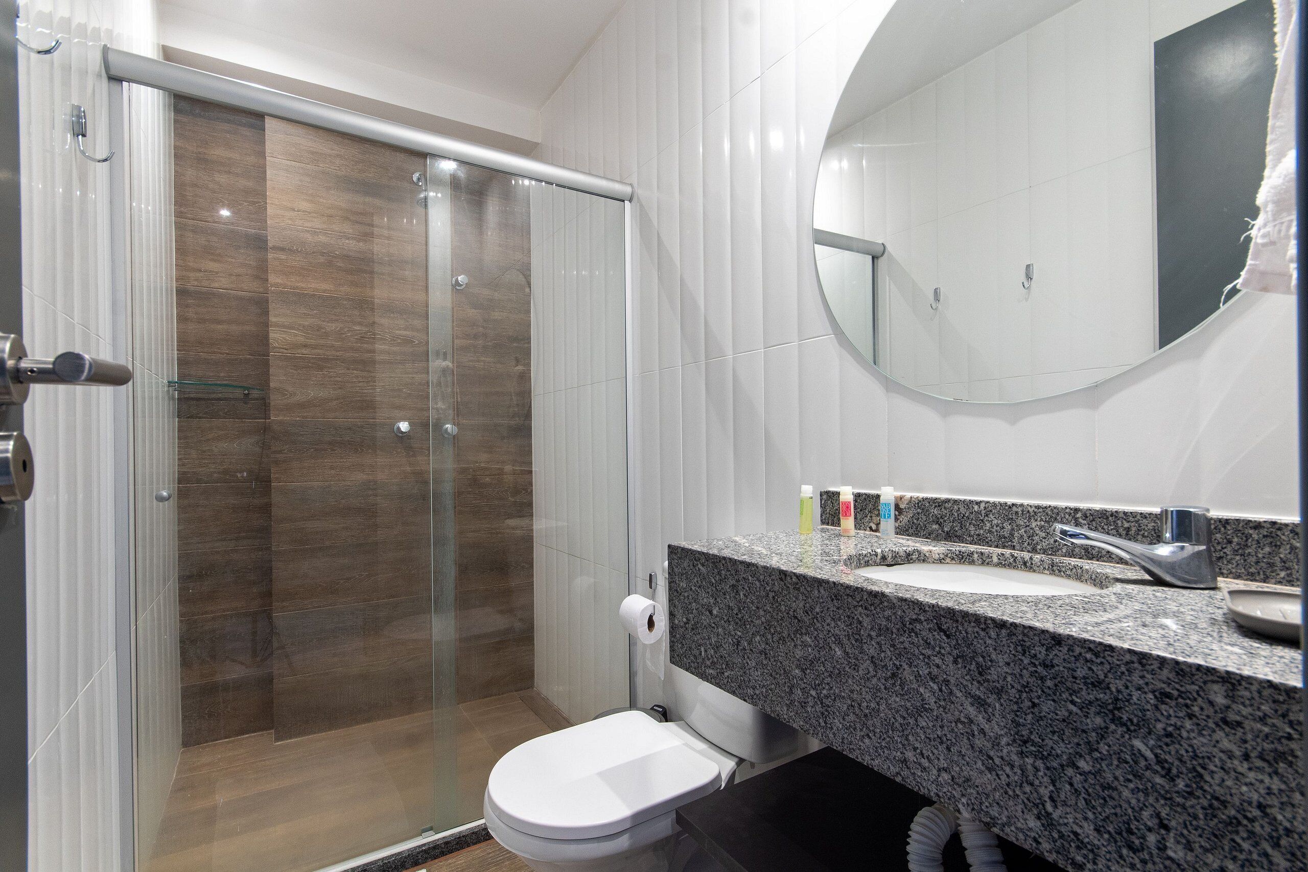 Un baño moderno con cabina de ducha de vidrio, inodoro blanco, lavamanos con encimera de granito y un espejo grande.