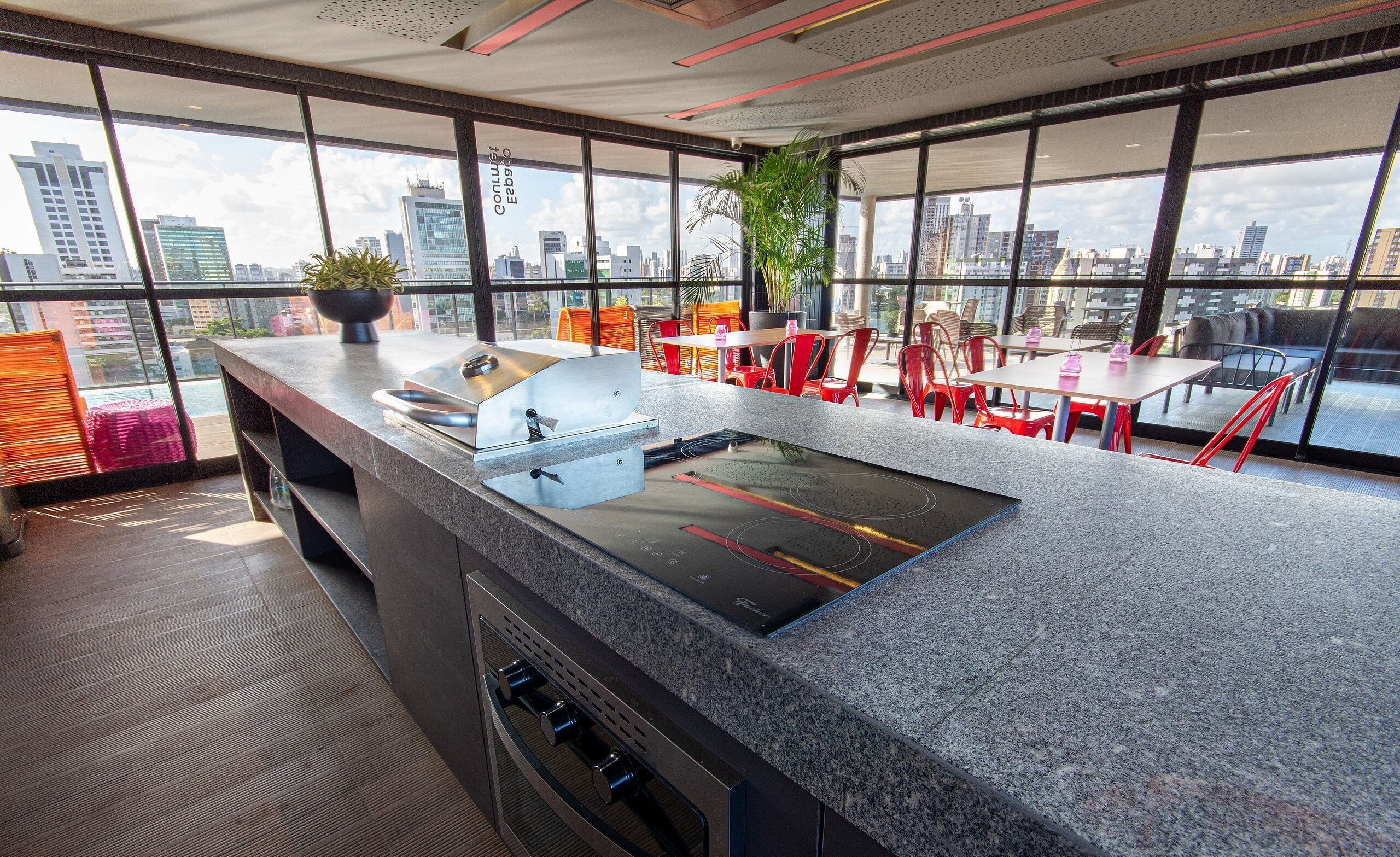 A modern kitchen with a built-in stove, a large window overlooking a cityscape, and red chairs around a table.