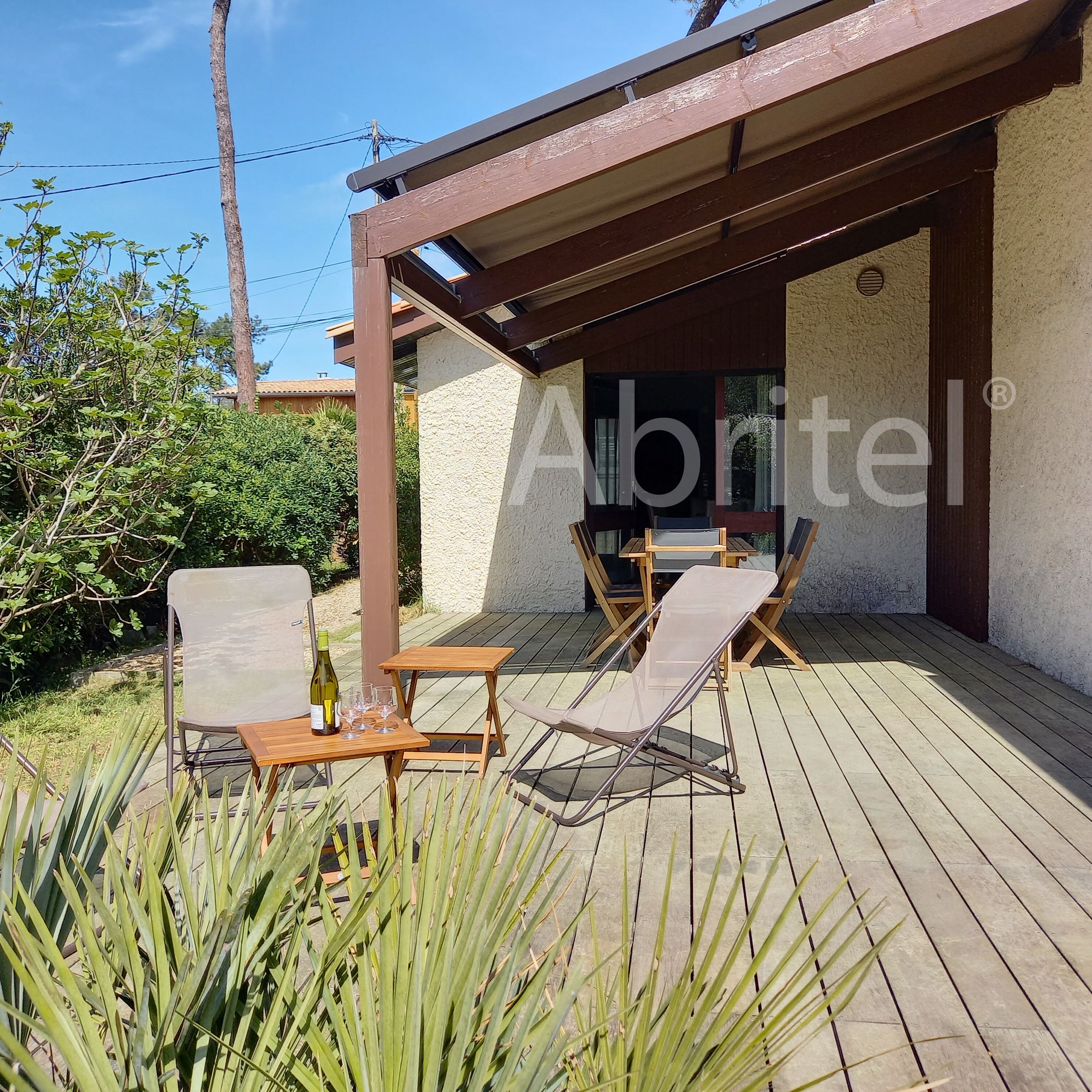 An outdoor patio with wooden furniture, a table with a bottle and glasses, and a view of a building with a window.