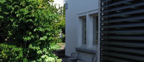 Ein Wohngebäude mit gepflasterter Terrasse, kleinem Garten und einem Fenster mit Rollläden.