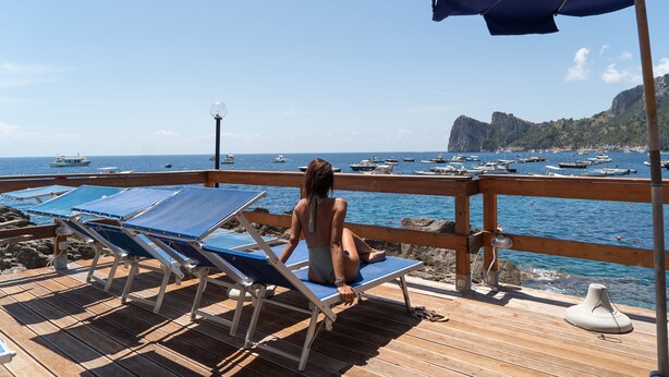 A wooden deck with blue lounge chairs and a large umbrella, overlooking a marina with boats and rocky cliffs.