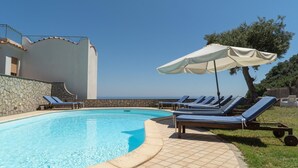 A swimming pool with lounge chairs and an umbrella, surrounded by stone walls and a clear view of the ocean.