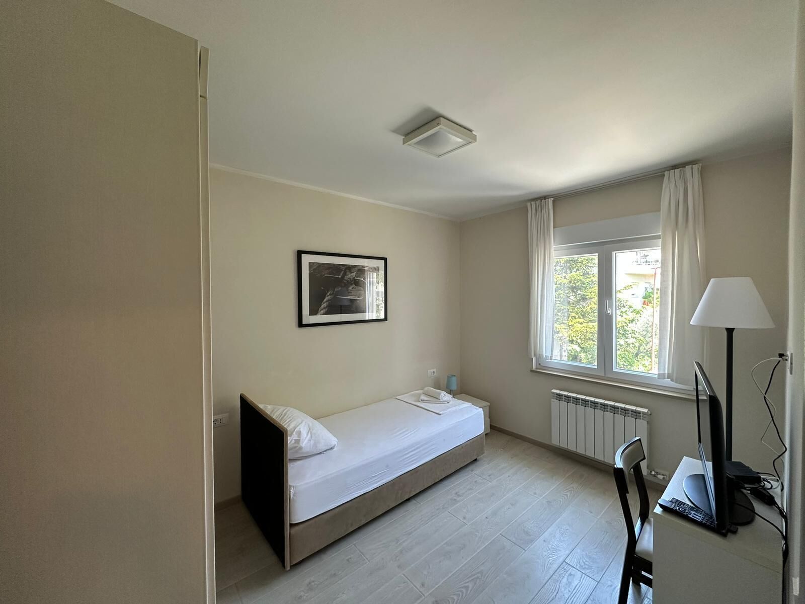 A small, minimalist room with a single bed, a desk with a computer, a window with curtains, and a radiator.