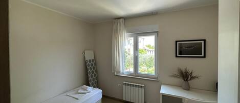 A small, well-lit room with a single bed, a radiator, a window with curtains, a desk with a chair, and a framed picture on the wall.
