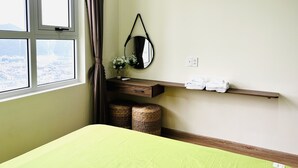 A hotel room with a bed, a window with curtains, a mirror, a shelf with towels, and a small wicker basket.
