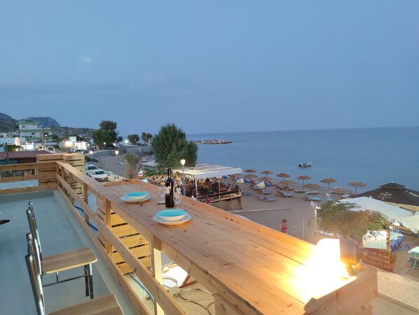 Una terraza de madera con mesas y sillas que da a una playa con sombrillas y vistas al mar.