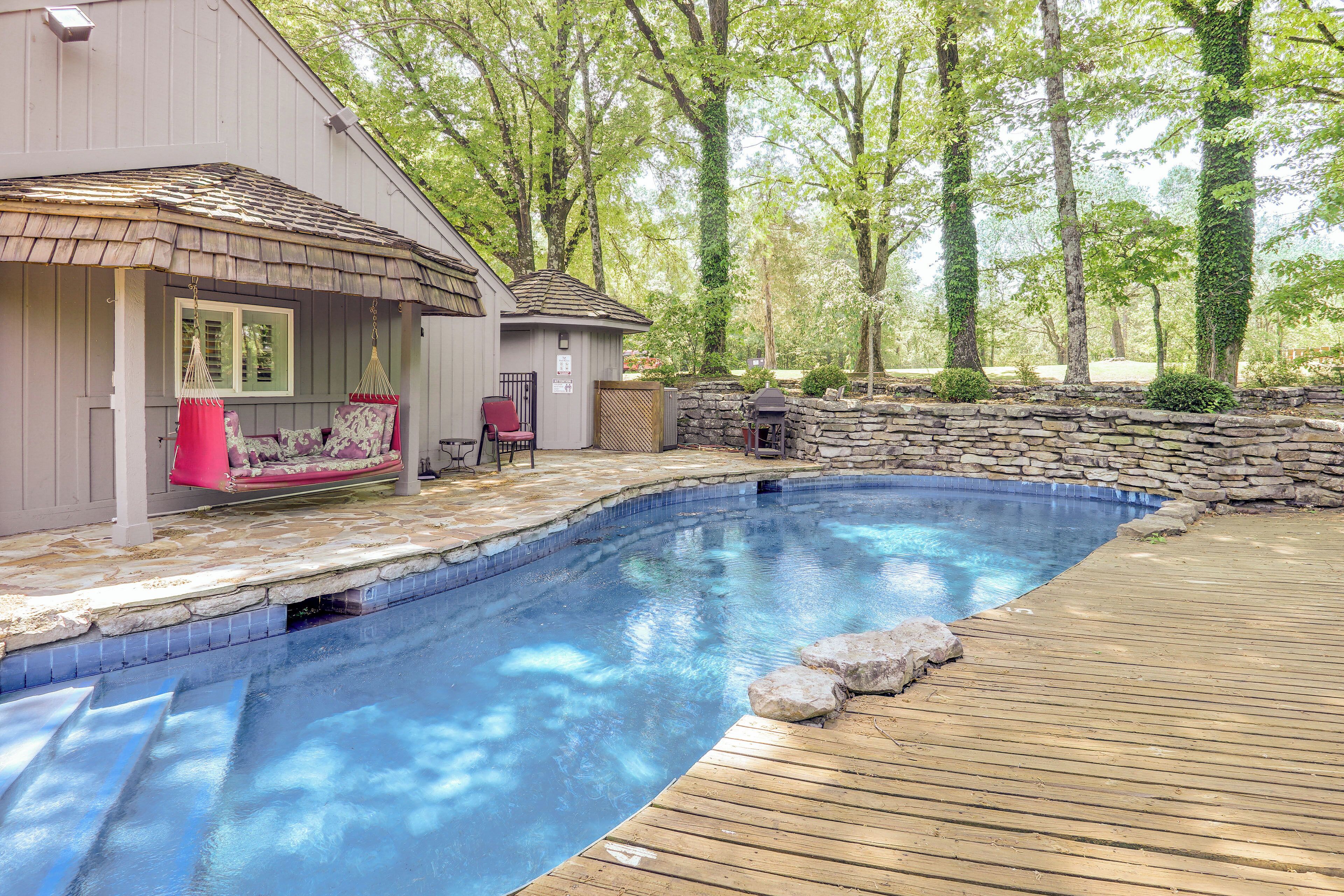 Une piscine avec une terrasse en bois, un espace salon couvert avec un fauteuil rouge et un mur en pierre.