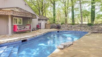 Une piscine avec une terrasse en bois, un espace salon couvert avec un fauteuil rouge et un mur en pierre.