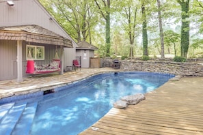 A swimming pool with a wooden deck, a covered seating area with a red chair, and a stone wall.