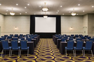 Un salón de conferencias con filas de sillas azules, una pantalla grande y candelabros.