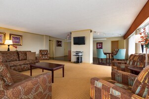 Habitación de hotel con sala de estar que incluye sofás estampados, una mesa de centro, un televisor de pantalla plana y un comedor con sillas.