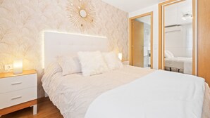 A bedroom with a large bed, a nightstand with a lamp, a wall-mounted mirror, and a door leading to a bathroom. - Los Ranchos 4-3 Apartment Levante Beach (Benidorm)