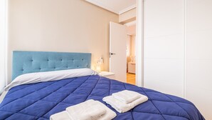 A neatly made bed with a blue quilt and white pillows, a white bedside table with a lamp, and a white door leading to another room. - Penthouse Ancla 562 Levante Beach (Benidorm)