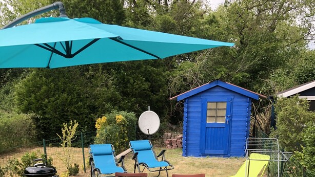 Een terras met een tafel en stoelen, een blauwe parasol en een blauwe schuur in een tuin.