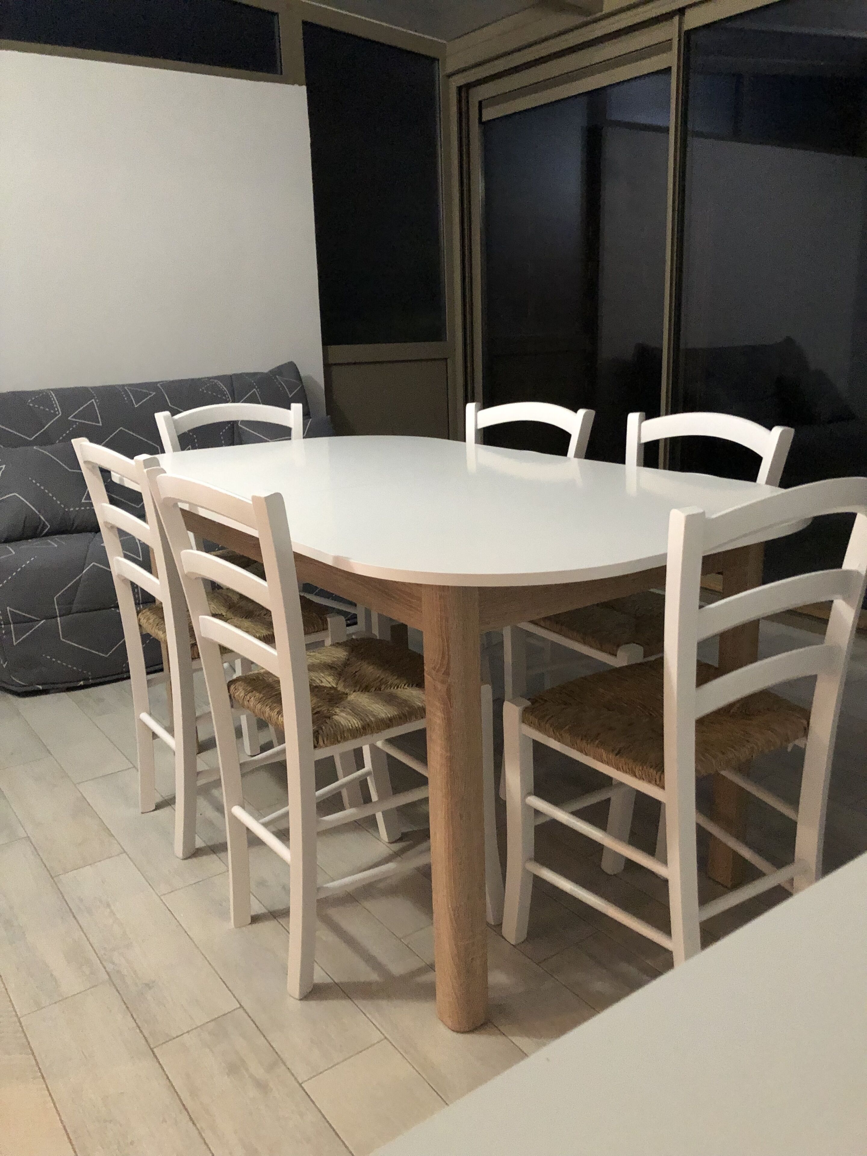 Une table de salle à manger ronde, entourée de chaises blanches, un canapé gris et une grande fenêtre.